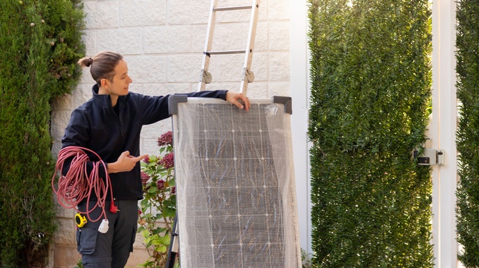 Eine Person in Arbeitskleidung hat ein langes Kabel um den rechten Arm gewickelt. Mit dem linken Arm hält sie eine verpackte Solarzelle. Neben der Solarzelle ist die Wand eines Gebäudes, an das eine Leiter angelehnt ist