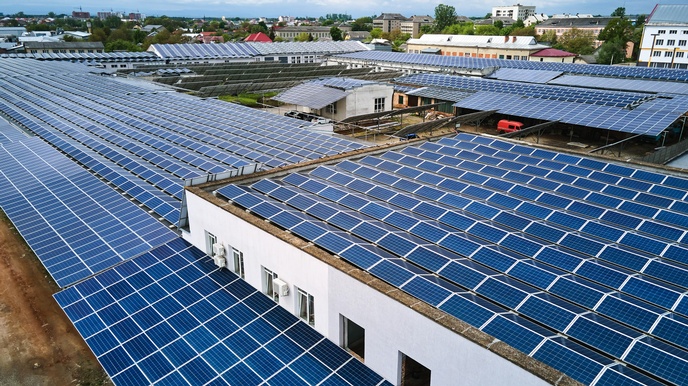 Hallen und Gerüste voller Solarpanele, im Hintergrund Stadtansicht unter bewölktem Himmel