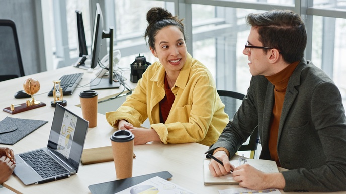 Zwei Personen an Arbeitstisch sitzend in Unterhaltung miteinander, am Tisch verschiedene Arbeitsutensilien, Dokumente mit statistischen Grafiken und Kaffeebecher