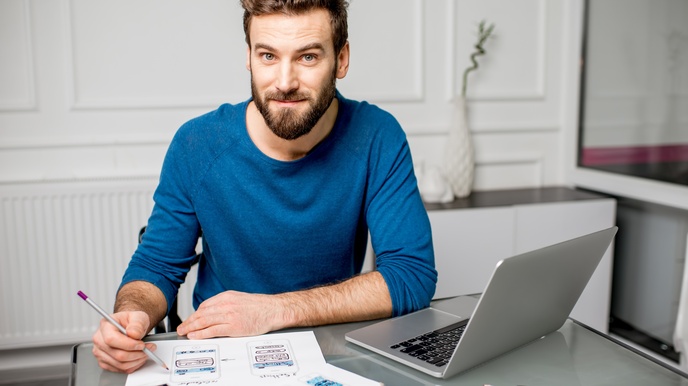 Person mit kurzen braunen Haaren,  Bart und blauem Pullover sitzt bei einem Schreibtisch mit Laptop und zeichnet mit Stiften ein UX Design auf Zetteln
