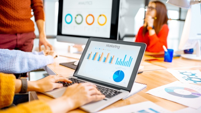 Auf einem großen Tisch steht ein aufgeklappter Laptop, auf dem eine Person tippt. Auf dem Bildschirm sind Diagramme und Statistiken mit der Überschrift Marketing. Am Tisch liegen weitere lose Zettel mit Statistiken