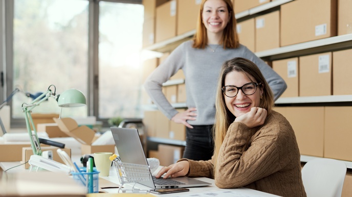 Zwei E-Commerce-Geschäftspersonen arbeiten im Büro. Auf dem Schreibtisch befinden sich ein Laptop, Kartons und Büromaterialien. Im Hintergrund sind Regale voller Kartons zu erkennen.