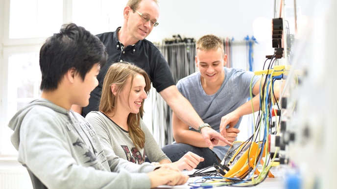 Seitliche Aufnahme von drei jungen Personen, die an einem Tisch sind. Vor ihnen sind verschiedene Kabel. Neben ihnen steht eine Person, die auf die Kabel deutet