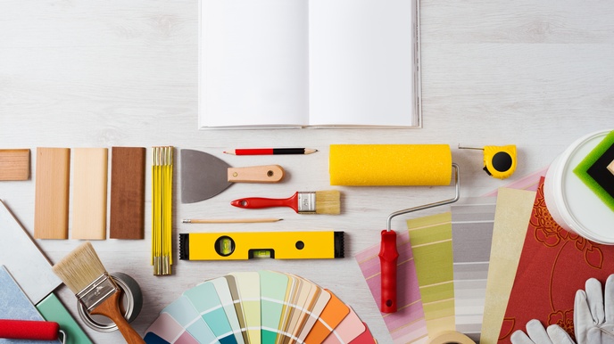 Verschiedene Utensilien fürs Malen und Tapezieren nebeneinander geschlichtet auf hellem Holzuntergrund: Pinsel, Stifte, Farbmuster, Wasserwaage, Spachtel, Klebeband und Farbrolle