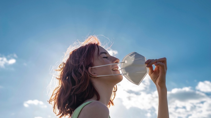 Lachende Person im Portrait in Seitenansicht vor sonnigem blauem Himmel mit Wolken zieht sich mit einer Hand Mundnasenschutz vom Gesicht