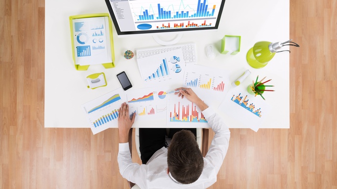 Person in Vogelperspektive an Schreibtisch sitzend auf dem Computer mit Statistik am Monitor steht, analysiert unterschiedliche Dokumente mit statistischen Diagrammen