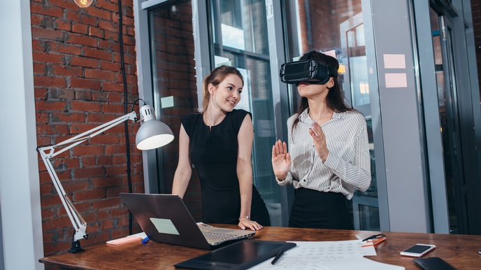 Eine Person mit Virtual Reality Brille steht an einem Schreibtisch während eine weitere Person neben ihr steht und diese beobachtet