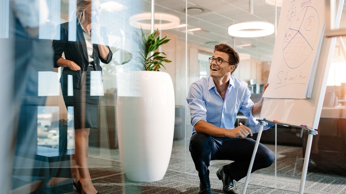 Person mit kurzen braunen Haaren, Brille und Businesskleidung kniet bei einem Flipchart mit Wording Startup, weitere Personen stehen daneben und blicken zu ihm in einem hellen, modernen Besprechungszimmer mit Glastüren