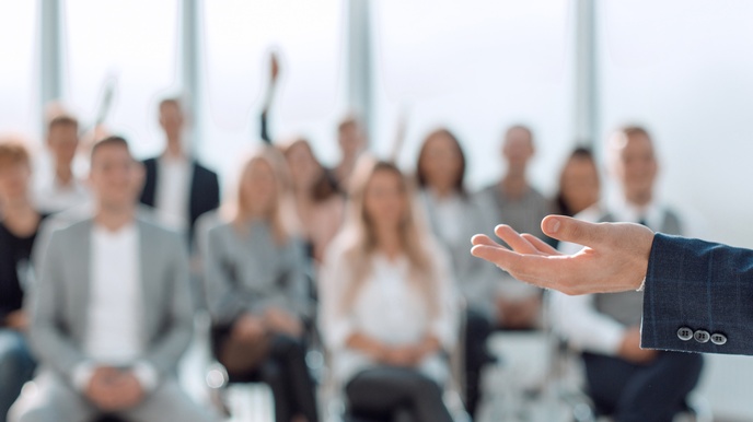 Hand mit blauem Sakko in Schärfe, im Hintergrund zeigt sich ein Publikum sitzend in der Unschärfe