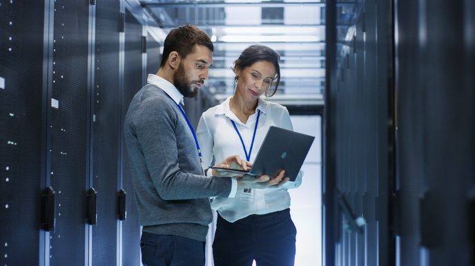 Zwei Personen stehen vor Servern und blicken auf aufgeklapptes Notebook, das eine Person in Händen hält