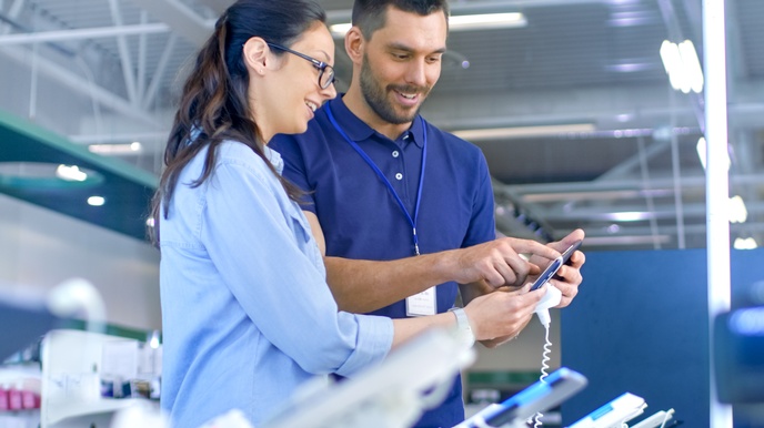 Seitliche Aufnahme zweier Personen, die nebeneinander in einem Geschäft stehen und ein Handy halten, das mit einem Kabel fixiert ist. Die rechte Person hat einen Ausweis um den Hals. Beide blicken auf das Handy