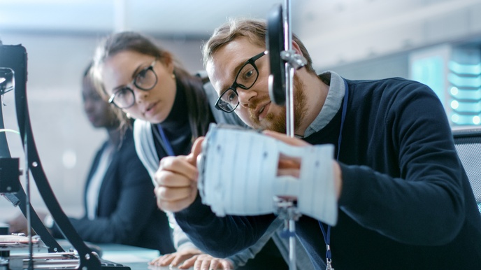 Zwei Personen mit Brillen blicken auf verschwommenes, 3D-gedrucktes Modell, im Hintergrund verschwommen weitere Person 