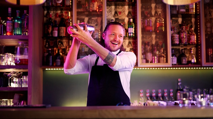 Person mit  schulterlangen nach hinten gefassten Haaren, Bart, weißem Hemd und dunkler Schürze steht hinter einer Theke vor einem Spirituosenregal und gießt eine Flüssigkeit von einer Spirituosenflasche in einen Cocktailshaker
