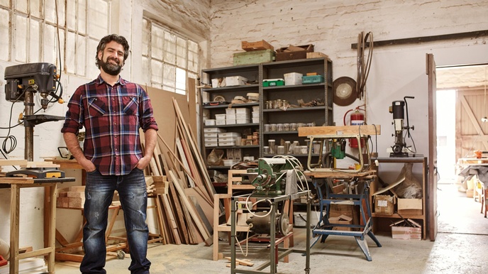 Cooler, lächelnder Tischler mit Bart steht in seiner Werkstatt mit Händen in Hosentaschen