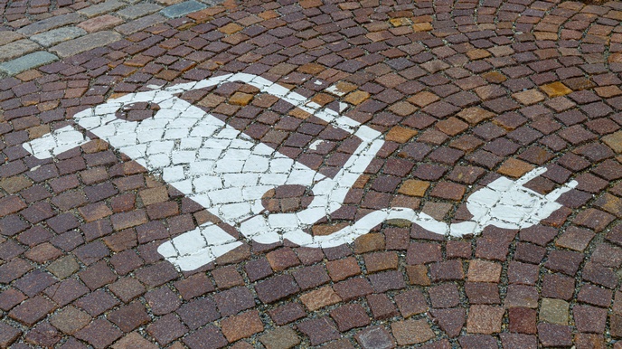 Auf einem Kopfsteinpflaster sieht man den Hinweis auf eine E-Lade-Station für ein Elektroauto als Bodenmarkierung in weißer Farbe. Das Symbol dominiert das Bild, das Kopfsteinpflaster ist nass vom Regenwetter. 
