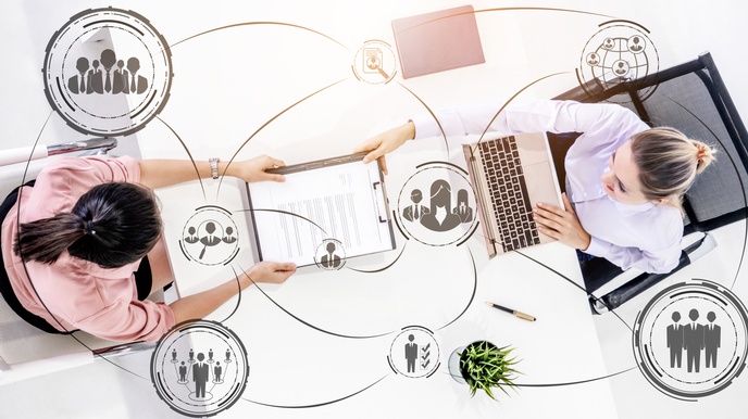 Zwei Personen sitzen sich an einem weißen Tisch gegenüber, die rechte tippt in einem Laptop, die linke hält ein Klemmbrett mit einem Dokument. Im Bild sind gezeichnete Linien, die zu Symbolen von Personen in Kreisen und einer Topfpflanze führen