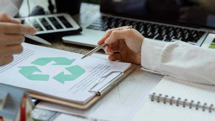 Auf einem Tisch liegt ein Klemmbrett mit einem Zettel, auf dem das Recyclingsymbol ist. Zwei Personen deuten jeweils mit einem Stift auf das Symbol
