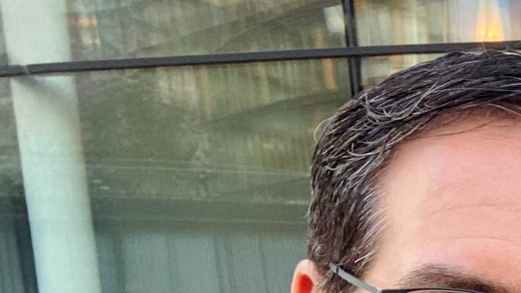 Porträt von zwei nebeneinander stehenden Personen vor einem Gebäude auf einem asphaltierten Platz. Die linke Person hat kurze, dunkle Haare und trägt Brille. Die rechte Person hat offene, helle Haare und trägt ein dunkles Oberteil