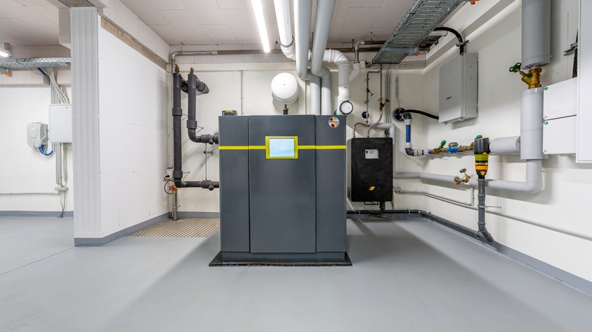 Ansicht einer Wärmepumpe: Dunkelgrauer metallischer Block mit gelber Linie und mittig platziertem Display in weißem Keller, ringsum Leitungen und Rohre verlaufend