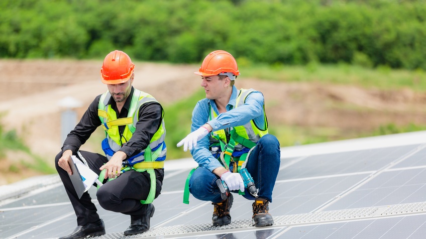 Zwei Personen in gelben Warnwesten mit orangen Schutzhelmen hocken auf Trittfläche auf Dach mit Solarpanelen und deuten auf bestimmte Stelle, im Hintergrund verschwommen belaubt Büsche