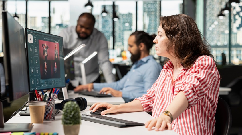Personen in einem Büro arbeiten an Computern, eine Person sitzt am Schreibtisch und nutzt Maus und Tastatur, zwei weitere Personen stehen und sitzen im Hintergrund.