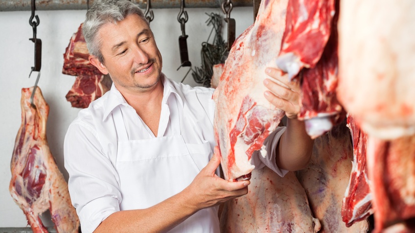 Person in weißer Schürze betrachtet großes Fleischstück, das zum Trocken aufgehängt ist, im Hintergrund an Haken weitere Fleischstücke