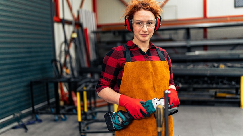 Porträt einer lächelnden Person mit Schutzbrille, Gehörschütz, Handschuhen, Schürze und geschlossenen Haaren, die in einer Werkstatt steht und ein Flexgerät hält