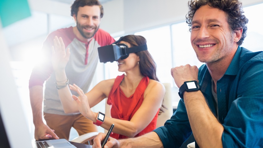 Drei lächelnde Personen an Schreibtisch stehend und sitzend, eine Person trägt VR-Brille und greift mit Händen in die Luft