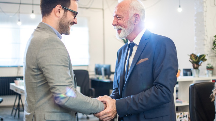 Zwei einander anlächelnde Personen in Anzügen schütteln sich die Hände, im Hintergrund verschwommen offener Büroraum