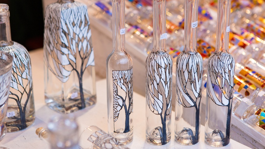 Nahaufnahme mehrerer leerer Flaschen in unterschiedlichen Formen mit hellen Stöpseln und kleinen Zetteln, auf denen Zahlen sind. Jede Flasche ist mit einem Baum mit weißen Blättern bemalt. Im Hintergrund liegen bunt bemalte Gläser