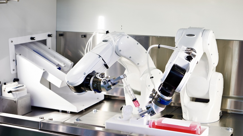 Zwei weiße Roboterarme stehen auf einer metallenen Oberfläche und halten gemeinsam eine medizinische Spritze, in der helle Flüssigkeit ist