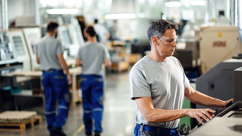 Seitenansicht einer Person in Arbeitskleidung mit Schutzbrille vor einer CNC-Maschine. Die Person schaut konzentriert auf den Bildschirm der Maschine. Im Hintergrund ist eine Industriehalle mit weiteren Arbeitenden Personen und Maschinen zu sehen.