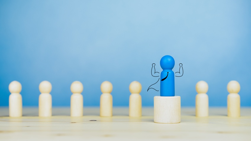 Kleine blaue Holzfigur steht auf Holzpodest, illustriert Krawatte, Umhang und erhobene starke Arme, im Hintergrund weitere naturfarbene Holzfiguren