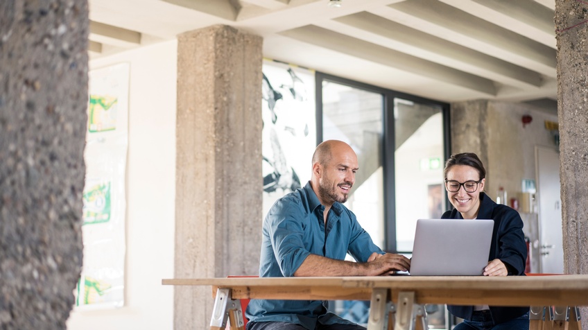 zwei Personen sitzen an einem Holztisch und schauen auf den Bildschirm eines Laptops