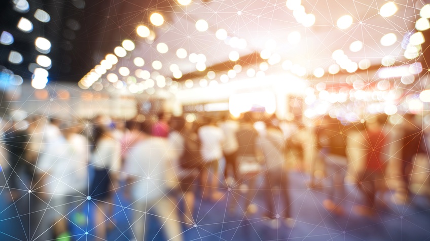 Verschwommene Aufnahme mehrerer Personen in einem Raum, die stehen oder sich bewegen. Darüber liegt eine Grafik, bestehend aus feinen, weißen Strichen, die kreuz und quer über das Bild verlaufen und miteinander verbunden sind