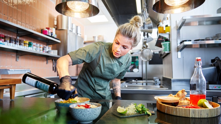 Person mit blonden geschlossenen Haaren und dunkelgrüner Arbeitsbekleidung steht in einer Gastronomieküche und bereitet Speisen zu