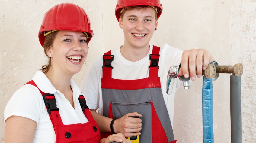 Zwei Personen in Arbeitskleidung stehen auf einer Baustelle im Innenbereich. Sie arbeiten an Sanitäranschlüssen. Die Personen halten ein Rohr und Arbeitsgerät in den Händen. Sie lachen fröhlich.
