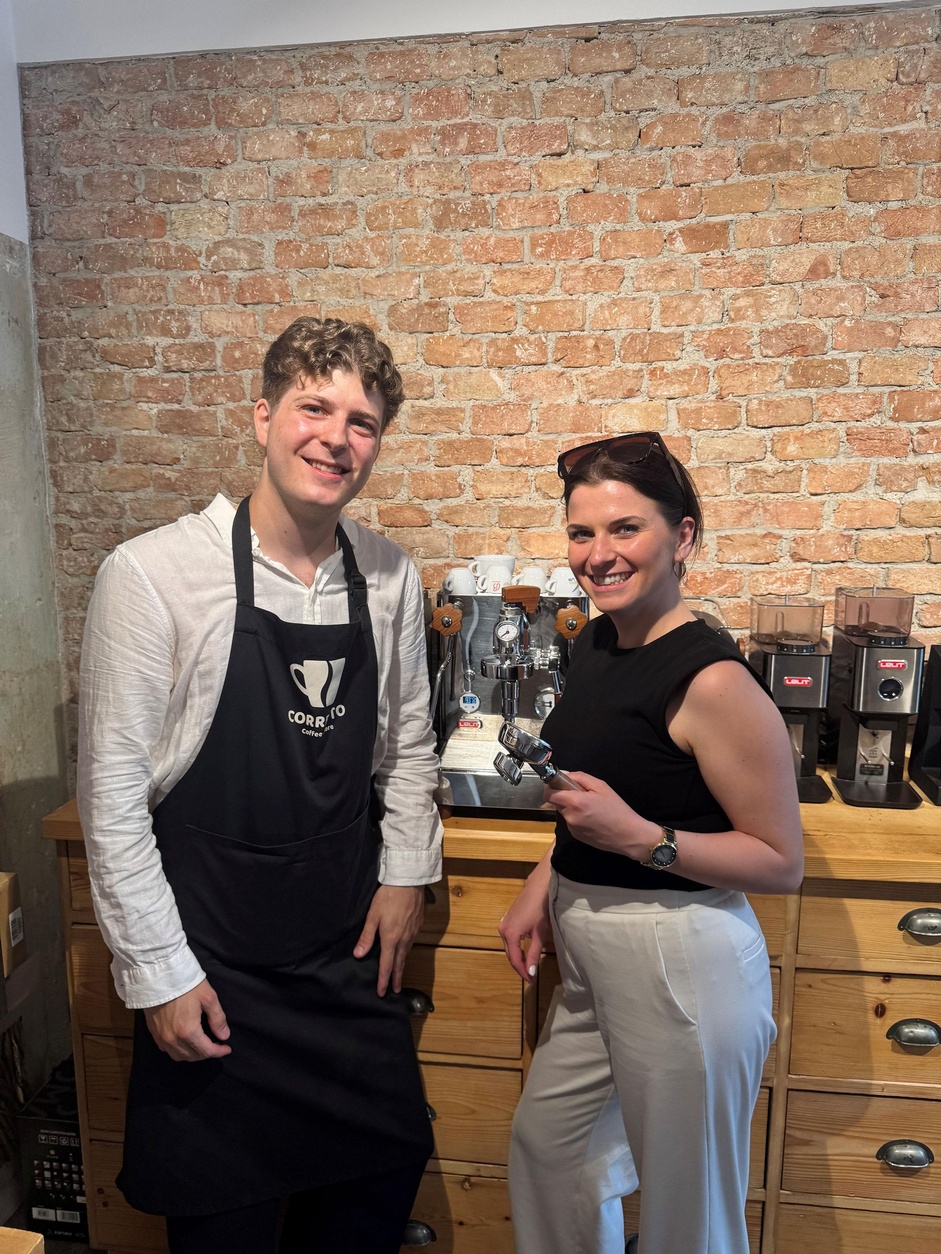 Zwei Personen stehen vor einer Espressomaschine und Kaffeemühlen in einer Kaffeerösterei oder Barista-Schulung. Eine Person trägt eine Schürze mit Corretto-Coffee-Logo, die andere hält einen Siebträger in der Hand. Im Hintergrund ist eine Backsteinwand zu sehen.