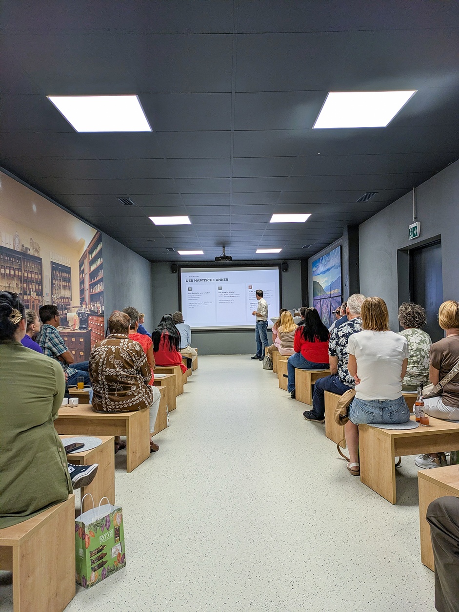 Teilnehmer:innen sitzen in einem Seminarraum auf Holzbänken und hören einer Präsentation zu, bei der vorne eine Person eine Folie zu einem wirtschaftlichen oder KI-bezogenen Thema erklärt.