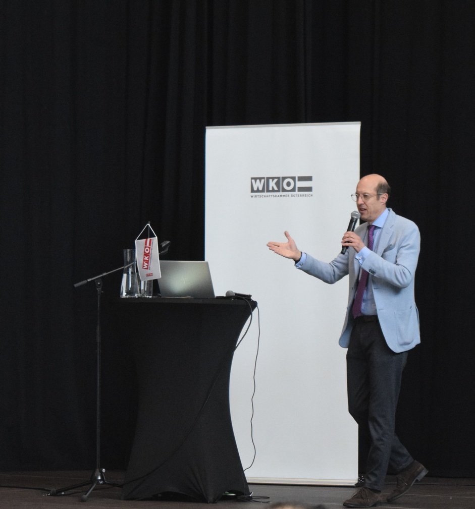 Eine Person steht neben einem Rundtisch, auf dem ein Laptop steht und ein Mikrofon liegt. Die Person spricht in ein Mikrofon. Hinter der Person ist ein weißer Banner, auf dem Wirtschaftskammer Österreich steht