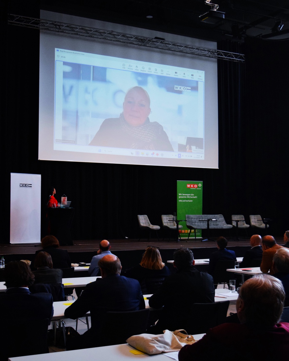 Rückenansicht mehrerer Personen, die in einem Raum an Tischen sitzen und alle zu einer Leinwand blicken, die an der Wand montiert ist und auf der eine Person zu den Personen spricht