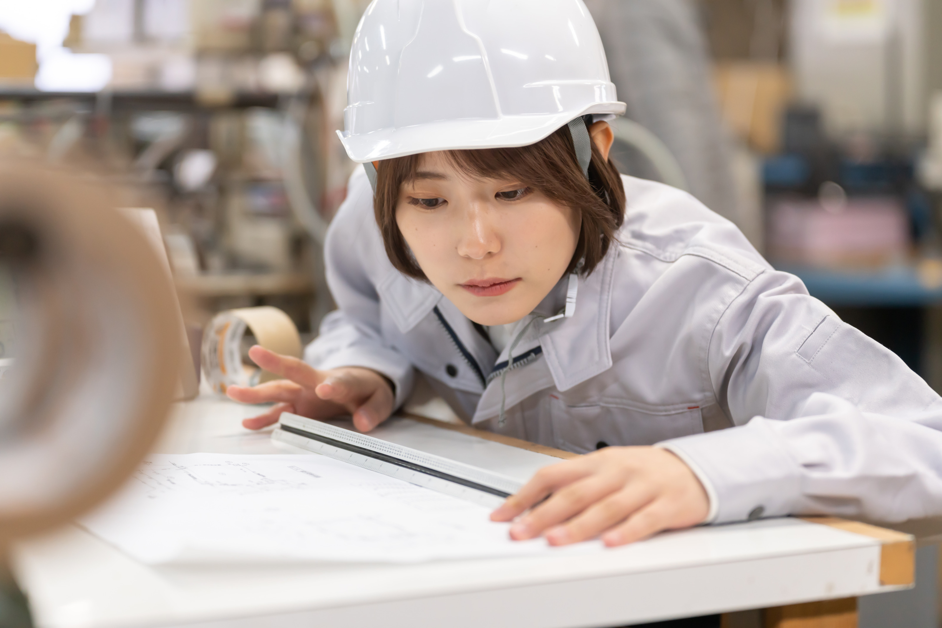 Person mit kurzen braunen Haaren, wei&szlig;em Schutzhelm und Arbeitskleidung ist &uuml;ber einen Tisch gebeugt, h&auml;lt ein Lineal &uuml;ber einen gezeichneten Plan und blickt fokussiert darauf, im Hintergrund zeigt sich in der Unsch&auml;rfe eine Werkst&auml;tte
