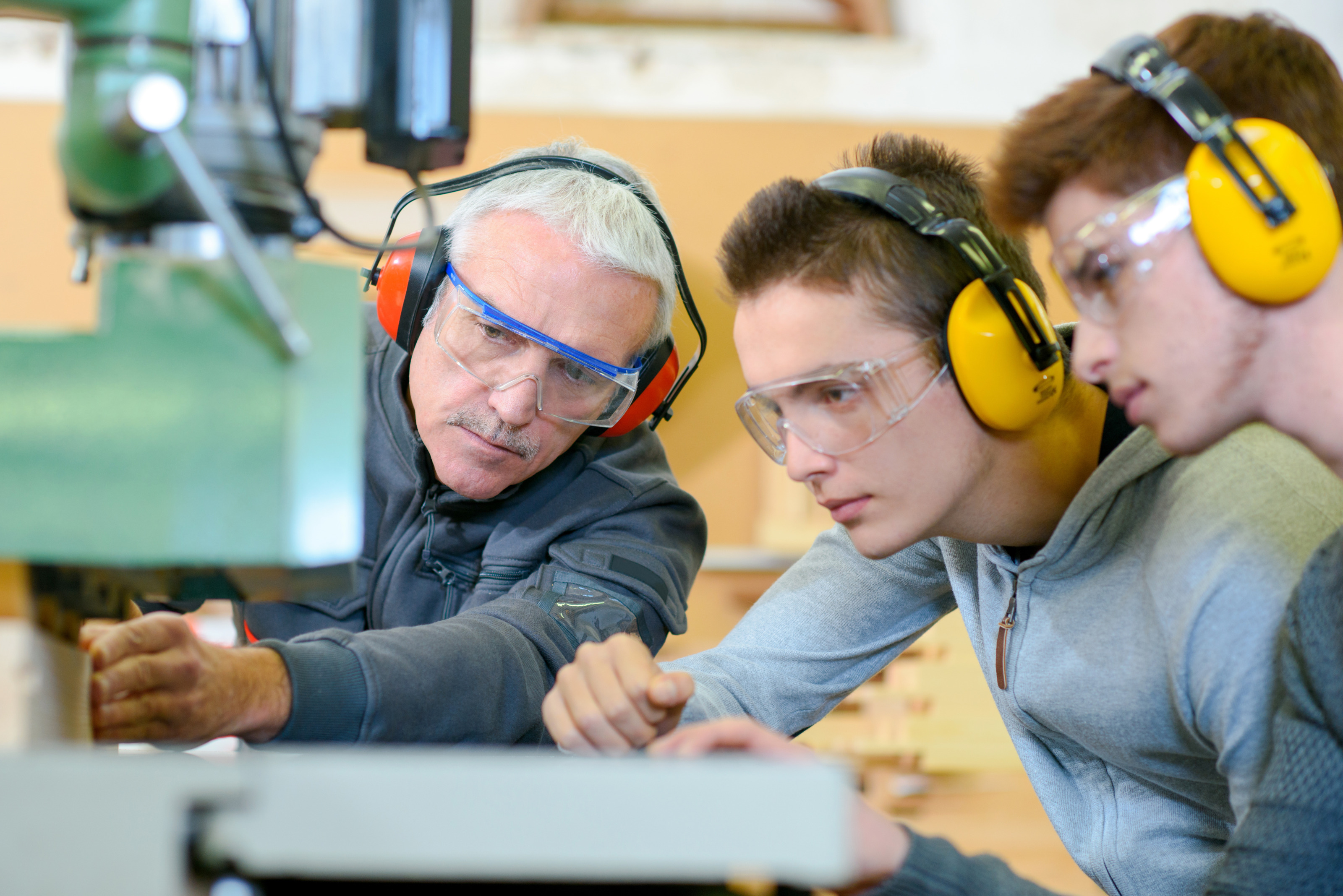 Drei Personen mit Schutzbrillen und Gehörschutz stehen an Werksmaschine, die eine der Personen bedient und blicken darauf