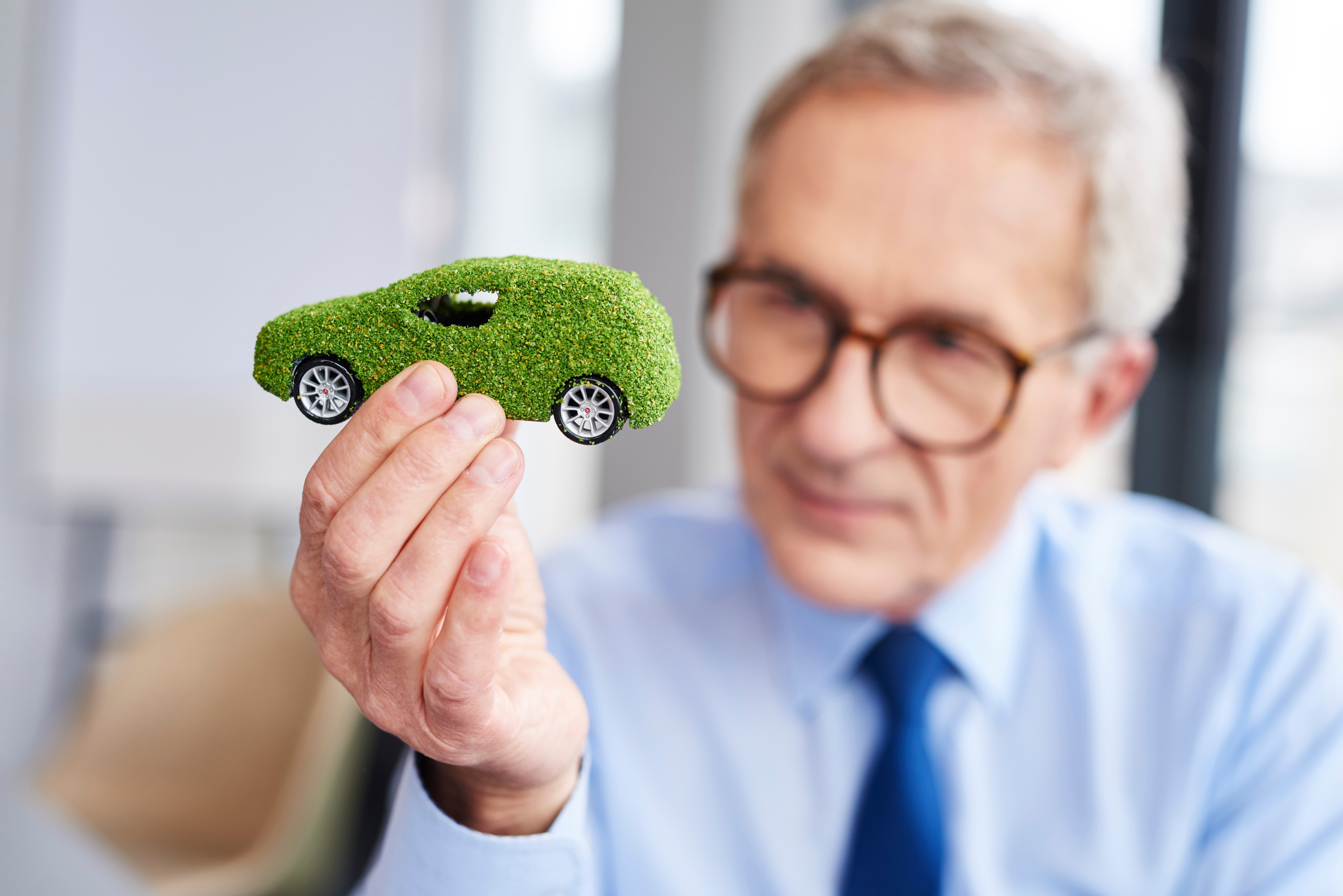 Eine Person mit Brille und blauem Hemd h&auml;lt ein kleines Miniaturauto in die Kamera. Das Auto ist mit gr&uuml;nen, unebenen Stoff &uuml;berzogen.