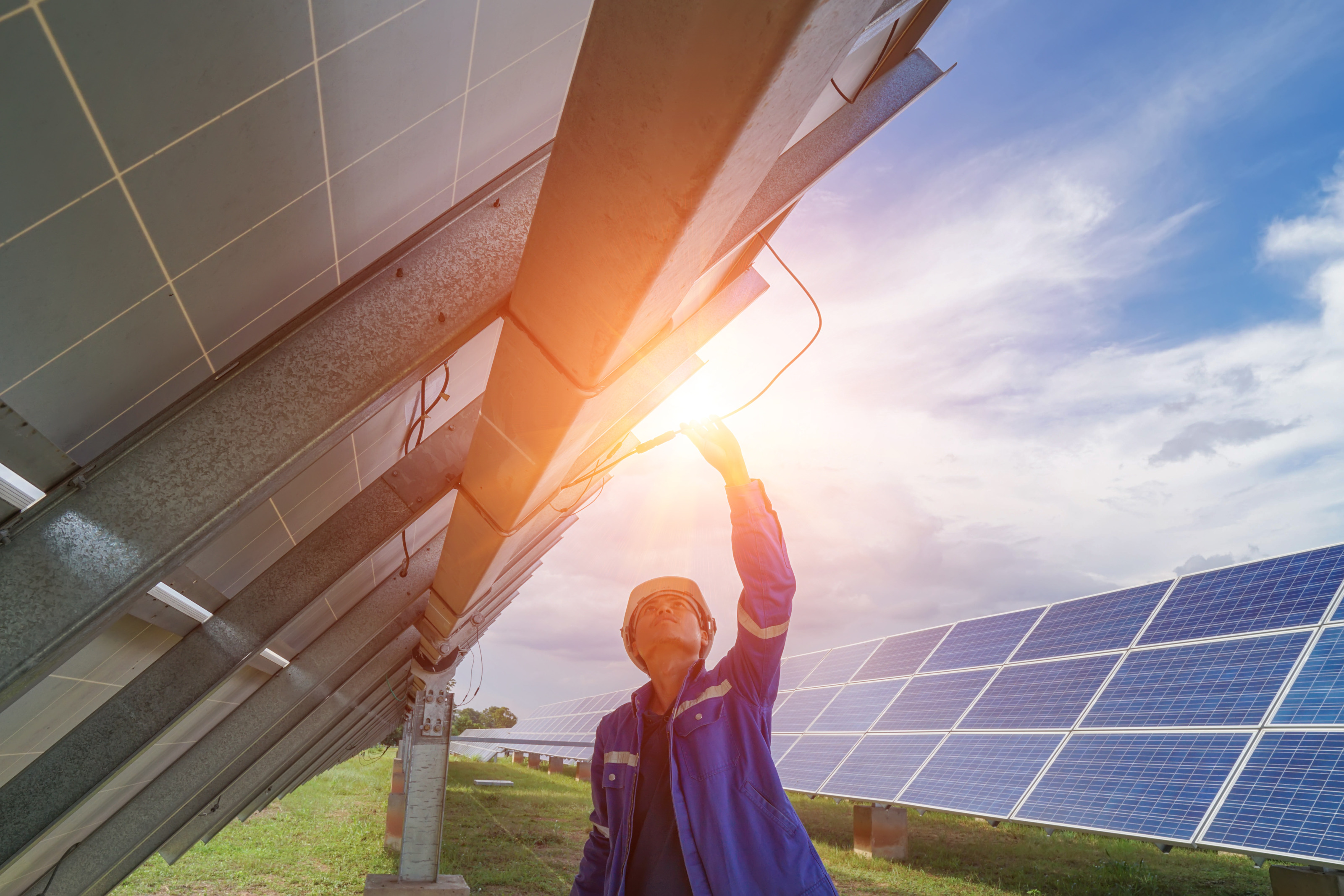 Person mit Helm streckt sich nach Oben und be&auml;ugt Kabel einer Solaranlage auf Metallger&uuml;st, im Hintergrund weitere Solarpanele auf gr&uuml;ner Wiese unter wolkenverhangenem Himmel im Sonnenuntergang