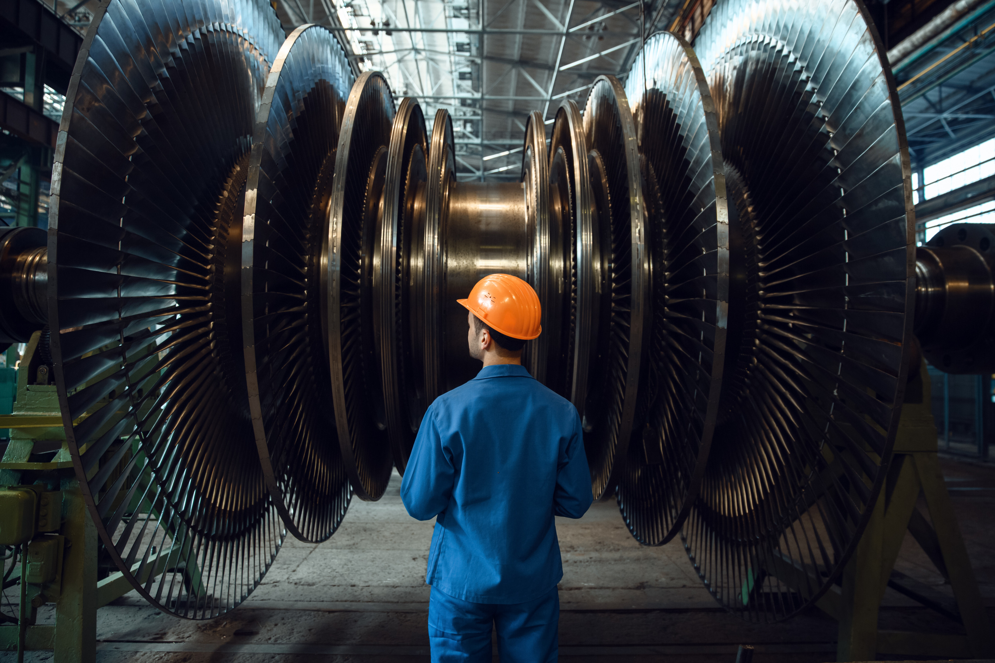 Person in blauem Overall mit orangem Helm in R&uuml;ckenansicht vor gro&szlig;er Turbine mit lamellenartig angeordneten Scheiben in Werkhalle stehend