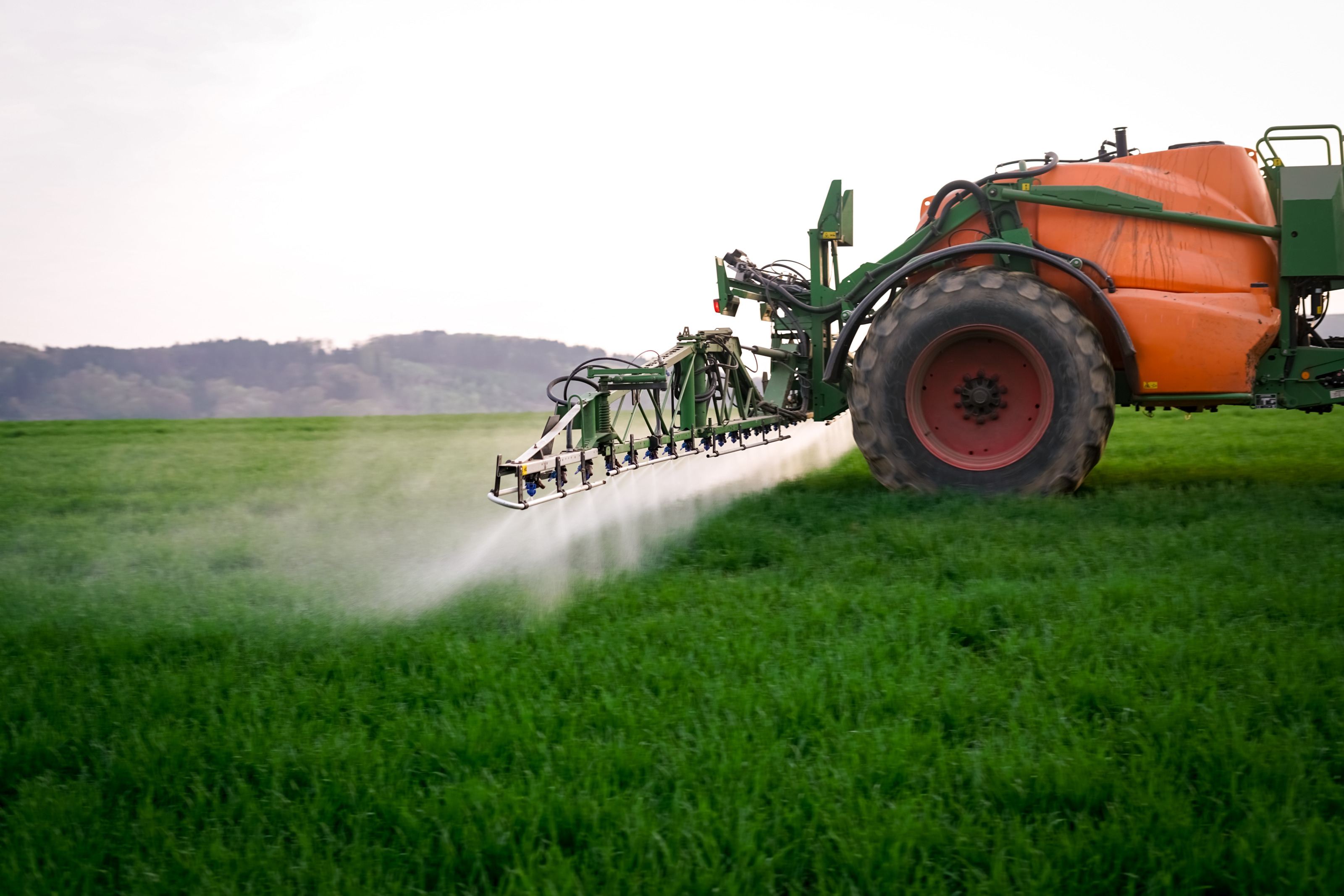 Orangegr&uuml;ne landwirtschaftliche Feldspritzmaschine im Ausschnitt f&auml;hrt &uuml;ber gr&uuml;n bewachsenen Acker und spr&uuml;ht aus aus D&uuml;sen am Heck Fl&uuml;ssigkeit auf darunterliegenden Boden, im Hintergrund verschwommen begr&uuml;nter H&uuml;gel unter grauem Himmel