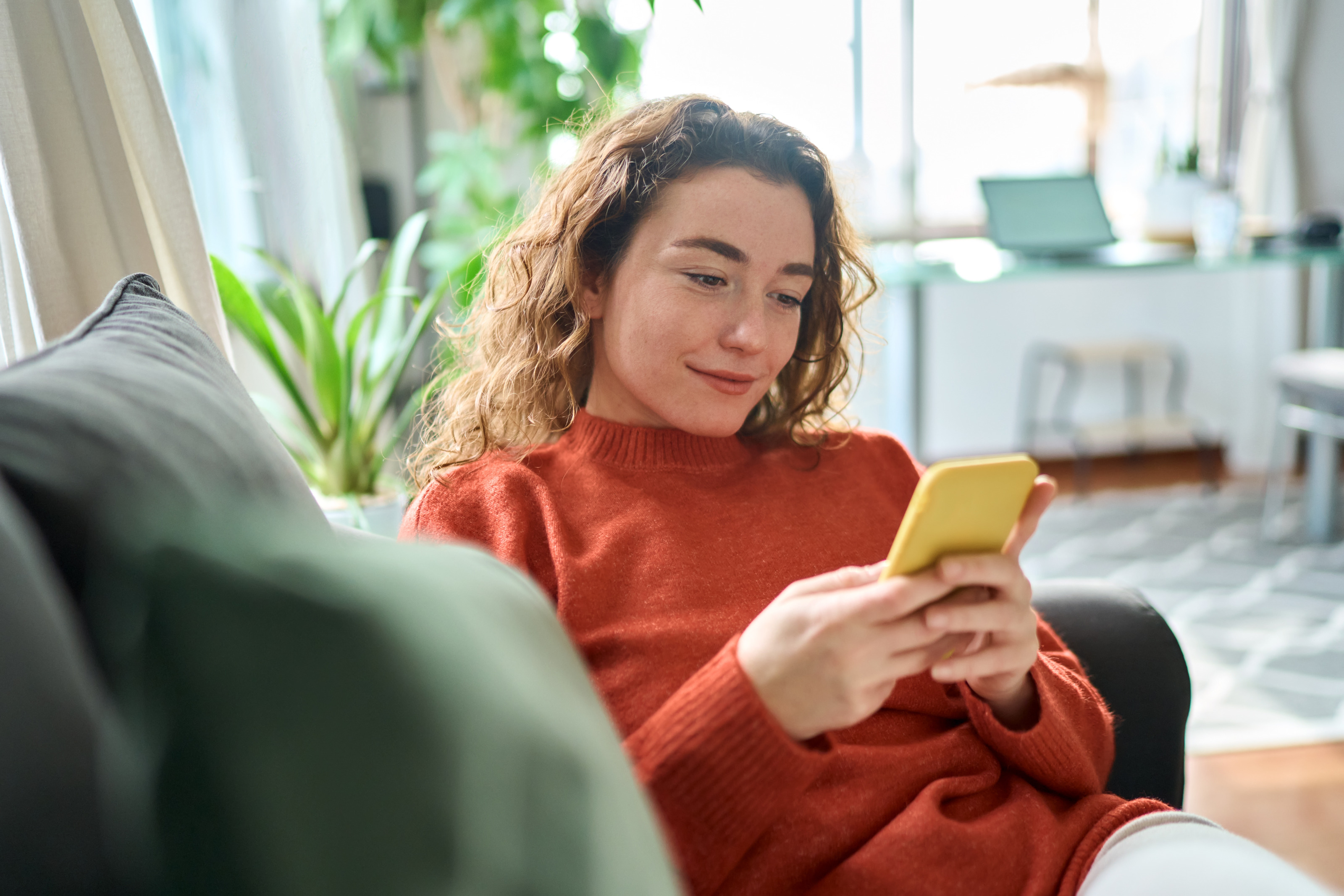 Eine Person mit lockigem Haar sitzt mit ausgestreckten Beinen auf einem Sofa in einem hellen Raum und blickt auf ein Smartphone, das sie mit beiden H&auml;nden h&auml;lt. Im Hintergrund sind Topfpflanzen und ein Schreibtisch