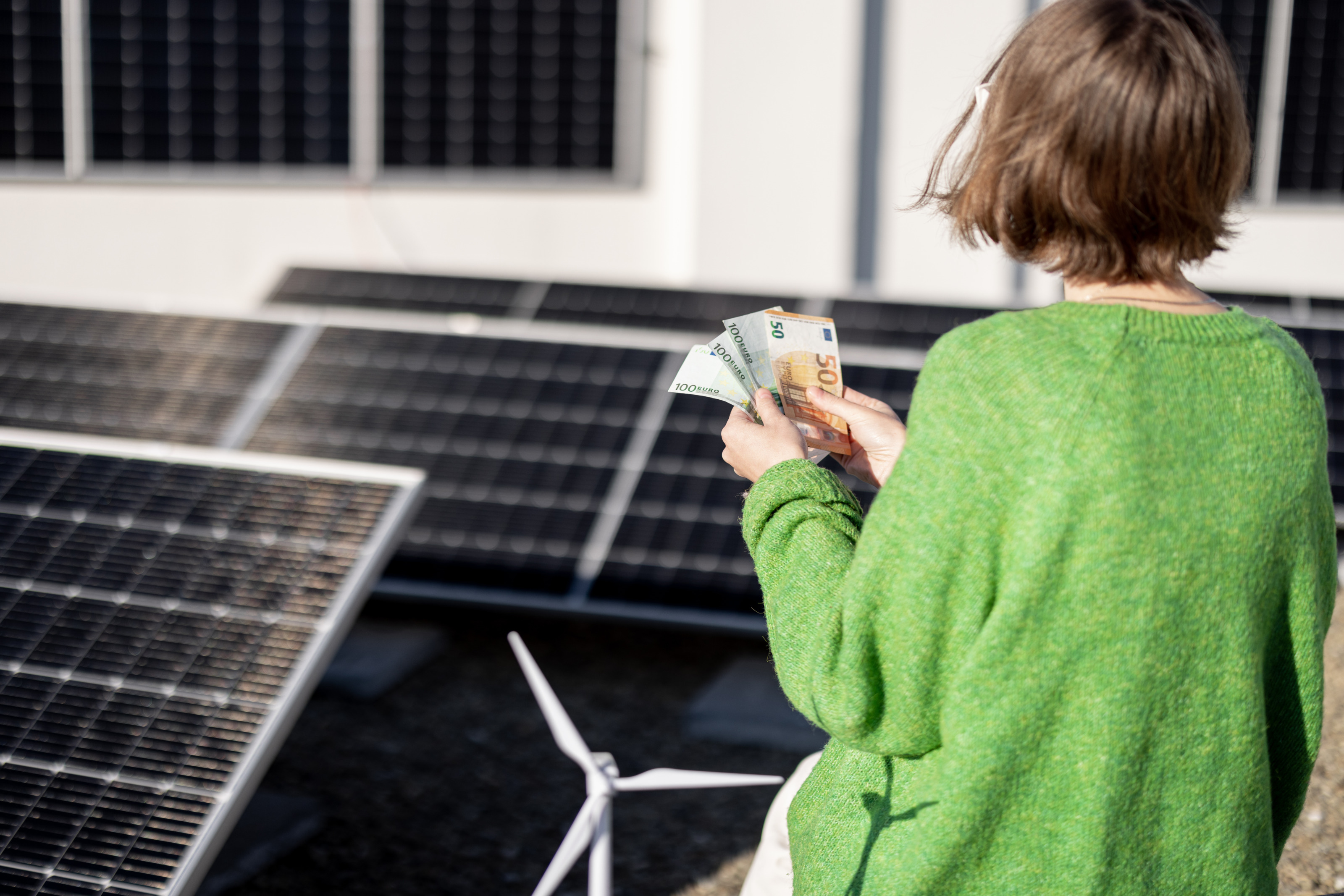 Person in gr&uuml;nem Pullover in R&uuml;ckenansicht steht inmitten einer Solaranlage und h&auml;lt F&auml;cher mit Eurogeldscheinen in H&auml;nden, im Vordergrund Modell eines Windrades