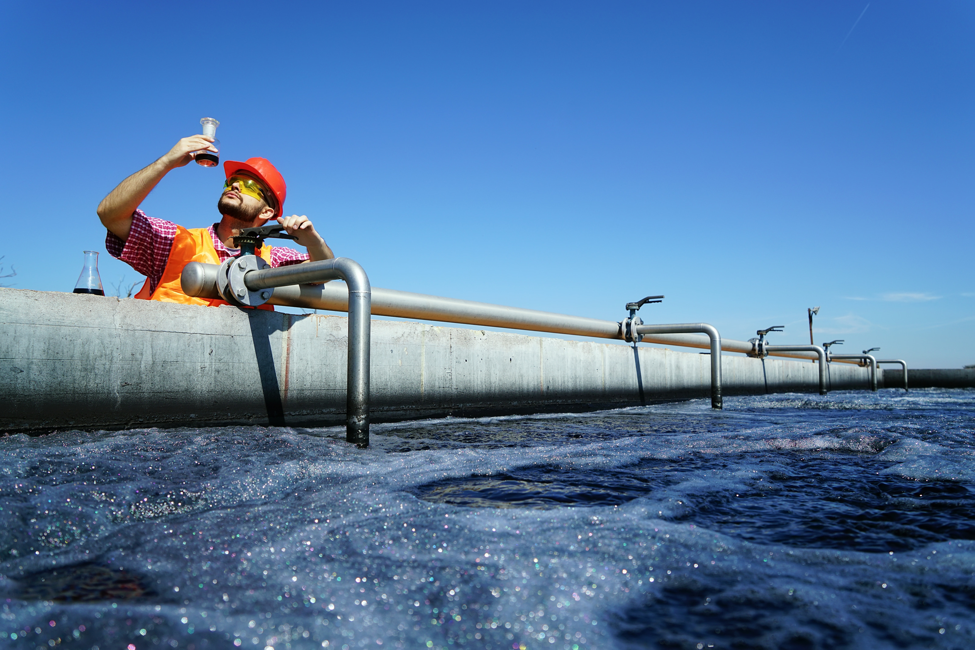 Person mit Helm steht an Wasseraufbereitungsanlage und blickt auf Testflasche mit Flüssigkeit